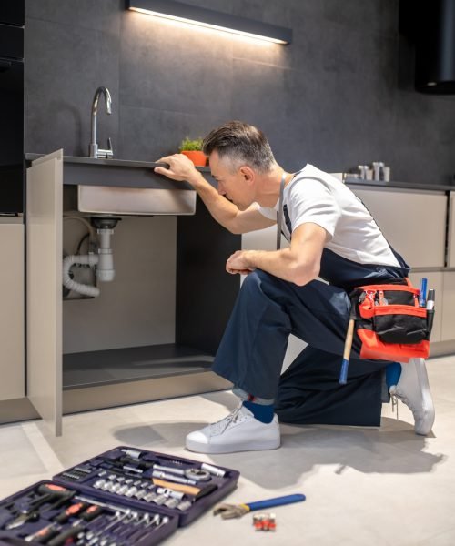 Household repair. Middle-aged man in blue overalls knelt down with hand touching sink carefully examining pipes in kitchen at home